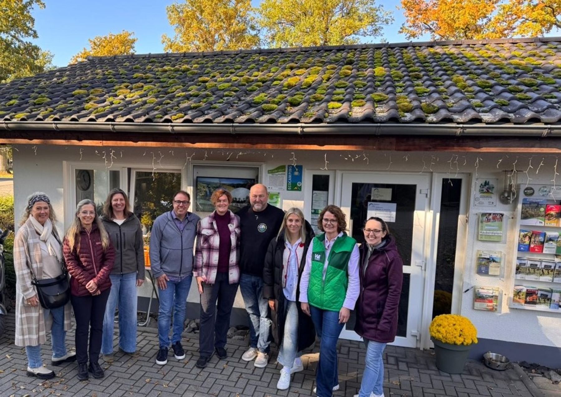 A group of ten people stands smiling in front of a small white building with a moss-covered roof. The building has brochures displayed outside, yellow flowers, and trees with autumn leaves in the background.